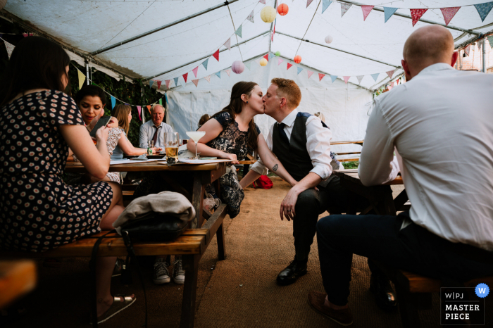 St. Albans, UK marriage reception party award-winning photo that has recorded Newlyweds kissing during the quiet tent reception - from the world's best wedding photography competitions offered by the WPJA