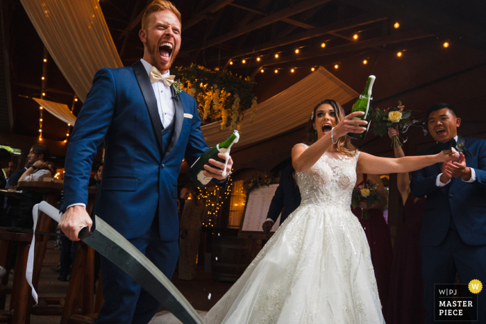 Blue Bridge Event Centre, Grawn, Michigan bekroonde foto van huwelijksreceptie waarop de bruid en bruidegom opgetogen zijn na het knallen van champagne met grote sabels - van 's werelds beste huwelijksfotografiewedstrijden aangeboden door de WPJA