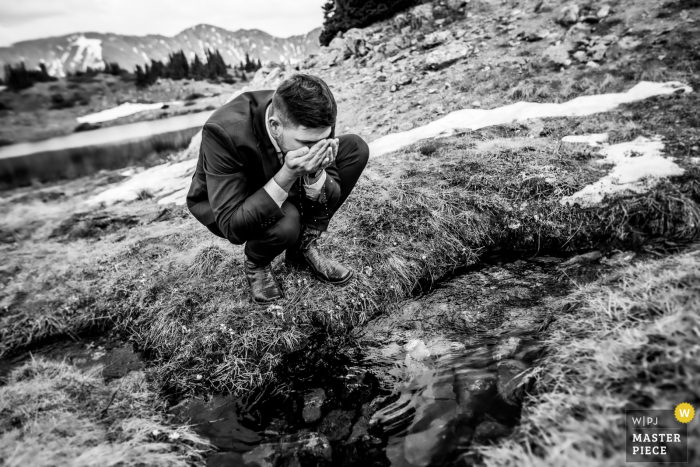 Loveland Pass, Colorado imagen ganadora del premio del día nupcial del novio bebiendo agua en un arroyo, de los mejores concursos de fotografía de bodas del mundo organizados por la WPJA