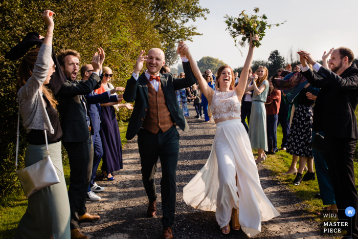 Ein dokumentarischer Hochzeitsfotograf bei Hoeve Engelendael hat dieses Bild eines glücklichen Brautpaares aufgenommen, das durch feiernde Gäste geht