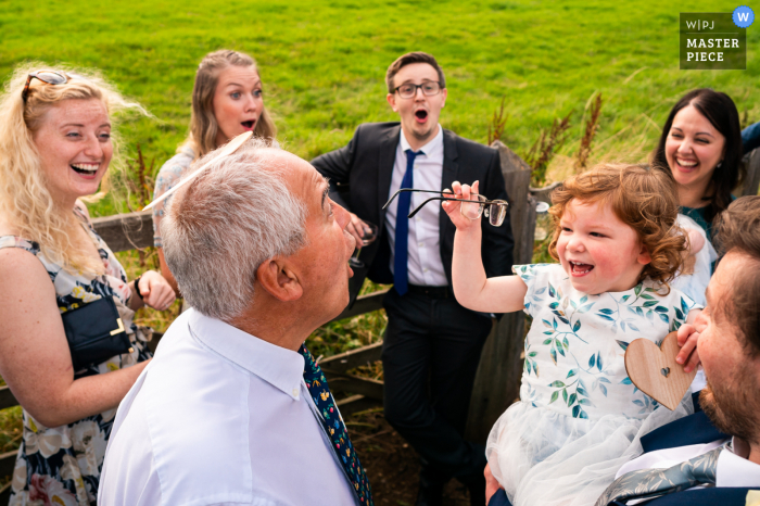 A top UK documentary wedding photographer at the Great Rollright Village Hall in Oxfordshire created this image of a Young child removing glasses from guests