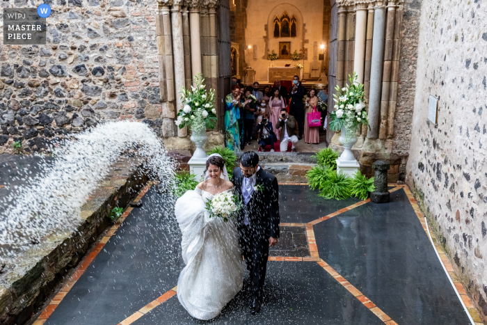 Een Italiaanse trouwfotojournalist bij Abbazia di Santa Maria di Maniace legde dit moment vast van deze echtgenoten die de kerk verlaten nadat ze hebben gewacht tot de regen stopte, wanneer er plotseling een regen van rijst op hen komt