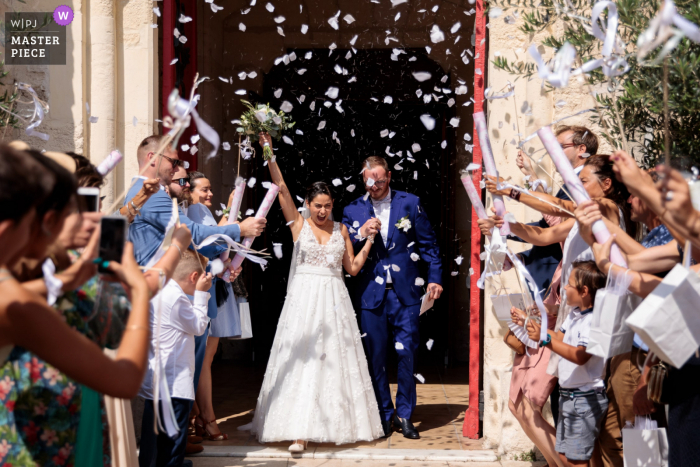 A top wedding photojournalist from Occitanie created this image as The bride and groom are leaving the church under confetti cannons 