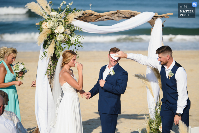 Sehen Sie sich diesen entscheidenden Moment im Sanderling Resort in Duck NC Hochzeitsbild an, das zeigt, wie der Trauzeuge bei der Strandzeremonie im Sand bei den Wellen Tränen und Schweiß für den Bräutigam abwischt