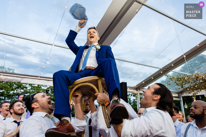 A top documentary wedding photographer in Wiscasset, Maine created this image as A groom is lifted in a chair during the hora