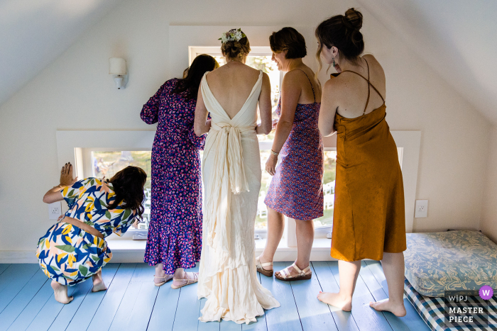 Um fotojornalista de casamento em Blue Hill, Maine, capturou este momento em A noiva e sua festa de casamento olhando pela janela enquanto os convidados entram na cerimônia