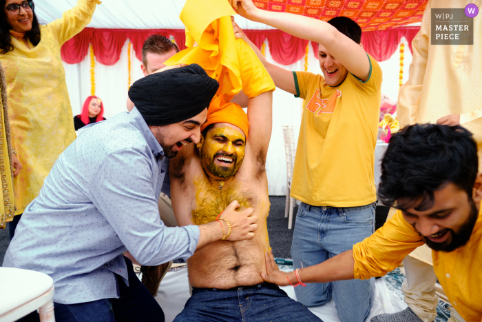 Melhor fotografia de casamento documental de Birmingham, Reino Unido, mostrando homens aplicando energicamente a pasta Haldi no noivo antes de seu casamento