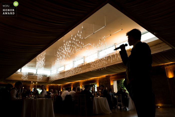 Een uitzonderlijke bruiloftsfoto uit Gloucestershire door een professionele Britse fotograaf bij Elmore Court met een silhouet van gasten die een toespraak houden op de locatie van de receptie