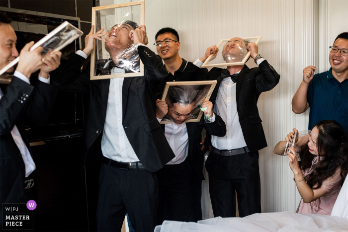 Imagem de casamento na China de um hotel em Sichuan dos irmãos noivos tentando perfurar o filme transparente em seus rostos enquanto as quatro pessoas segurando molduras de madeira da esquerda para a direita têm expressões diferentes