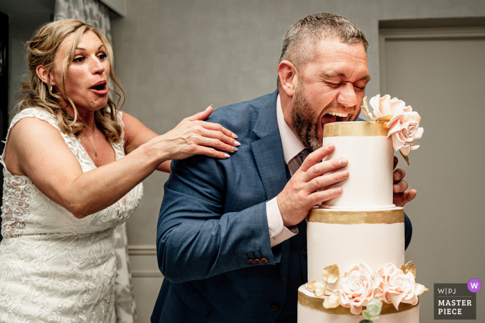 Beste grappige huwelijksfotografie van de locatie van het Wrightington Hotel in Greater Manchester, VK, waarop de bruidegom te zien is die in de cake bijt tijdens het aansnijden van de cake