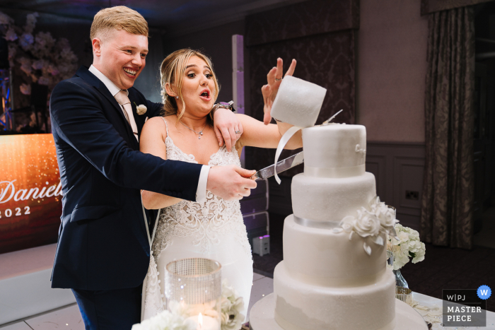 Beste fotografie voor de trouwlocatie in Manchester in de Mottram Hall, waarbij bruid en bruidegom worden vastgelegd die cake snijden en de bovenkant valt eraf