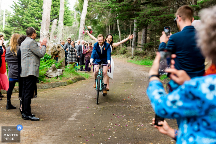 Best wedding photography from a Fort Bragg venue, Holly's Ocean Meadow in California capturing A bride and groom ride through a crowd of their guests on a tandem bicycle