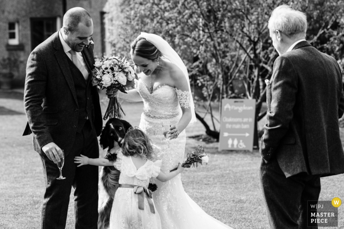 Fotógrafo de casamento documental no local do evento Pangdean Old Barn em West Sussex, Inglaterra, capturou uma menina de flores e um cachorro abraçados perto da noiva e do noivo após a cerimônia de casamento