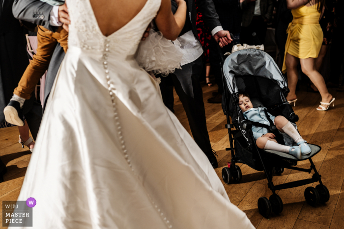 Documentary photography from Hodsock Priory wedding venue in Nottinghamshire from the dance floor, of a young child sleeping in his pram as his dad holds on as the bride dances in front of them