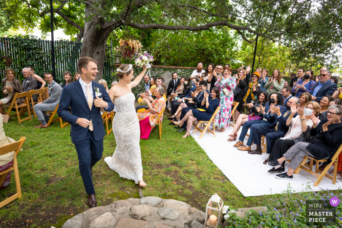Imagem de casamento de Altadena de uma cerimônia no quintal criada enquanto o casal recém-casado recua o corredor da cerimônia enquanto os convidados torcem Imagem de casamento de Altadena de uma cerimônia no quintal criada enquanto o casal recém-casado recua o corredor da cerimônia enquanto os convidados torcem