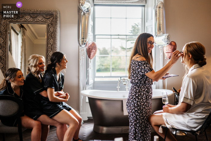 Der dokumentarische Hochzeitsfotograf am Veranstaltungsort Saltmarshe Hall in Yorkshire, Großbritannien, hat die Braut beim Schminken festgehalten, während ihre Brautjungfern zusehen, wie sie übereinander sitzen und mitlächeln, genau wie der Maskenbildner