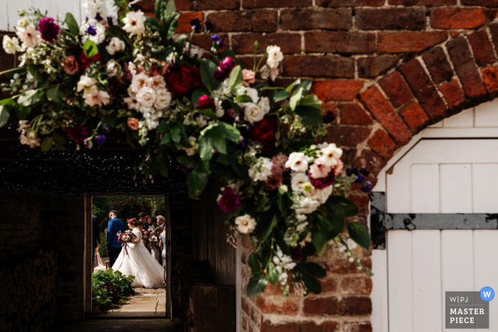 Fotografia de casamento de Brandesburton em Yorkshire, Inglaterra, de A noiva vislumbrada através de um arco floral lindamente adornado em seu local de casamento na fazenda