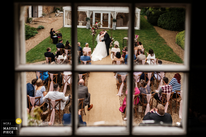 Photographie du lieu de mariage du Yorkshire à la Barff House exposant la mariée et le marié partageant un premier baiser avec leurs invités de mariage qui regardent, capturés depuis une fenêtre à proximité