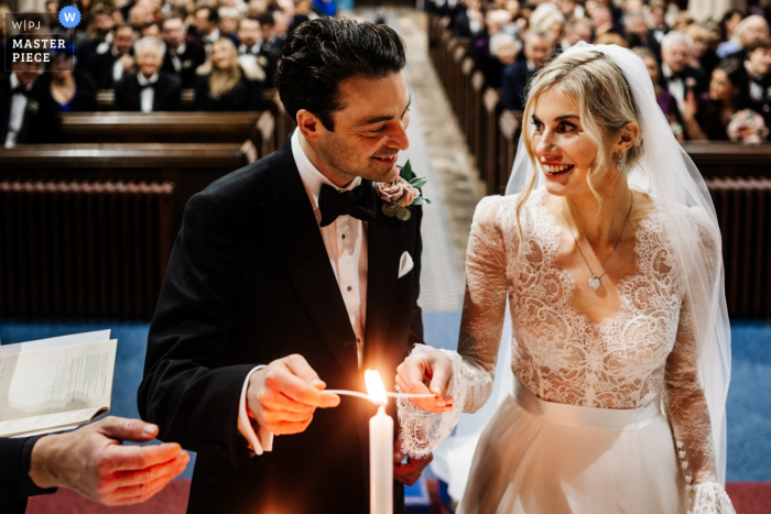 Fotografía documental de una boda en la Iglesia del Castillo de Berkeley en Gloucestershire que muestra a la novia y el novio sonriendo mientras encienden una vela juntos en la ceremonia de su boda, con la mano de su vicario dirigiéndolos visto en la esquina