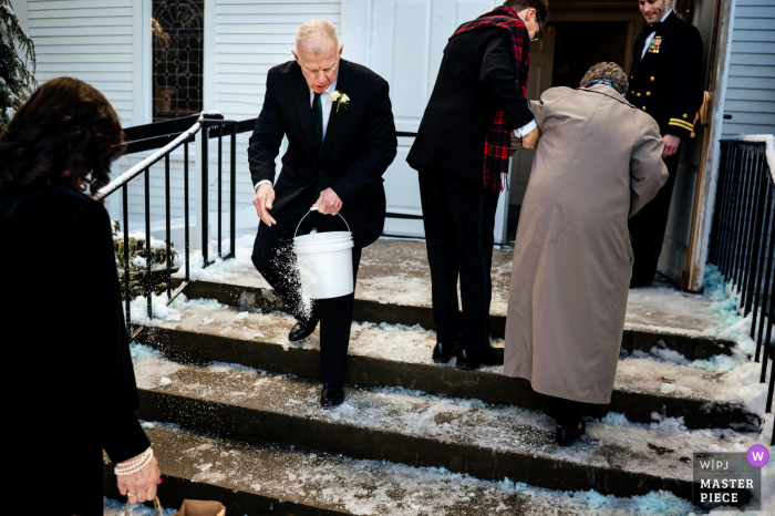 Beste huwelijksfotografie van The Equinox Resort in Vermont, gemaakt terwijl de vader van de bruidegom zout op de treden strooit om te voorkomen dat gasten op het ijs uitglijden