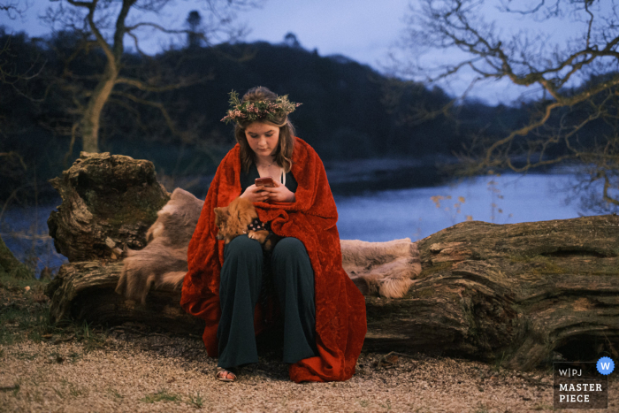 Fotografia documental do local de casamento Finnebrogue Woods em County Down, Irlanda do Norte mostrando a dama de honra mais jovem sentada sozinha acima do lago durante a recepção noturna, acompanhada por um cachorro pequeno