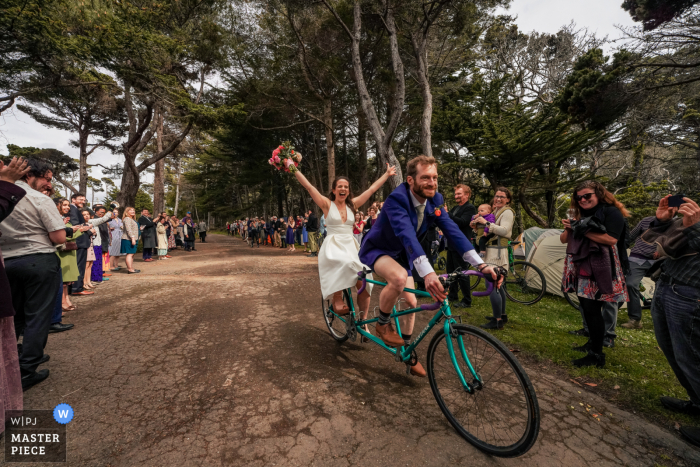 Documentaire trouwfotograaf op Holly's Ocean Meadow-evenementenlocatie in Fort Bragg, Californië legde dit vast. De bruid en bruidegom rijden hun receptie binnen op een tandemfiets voor hun juichende gasten