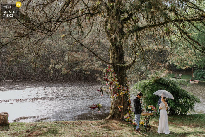 Fotografia dokumentalna z miejsca ślubu, Hotel Parador Hampel, w Rio Grande do Sul, przedstawiająca Parę Młodych w Deszczu podczas ceremonii plenerowej nad wodą