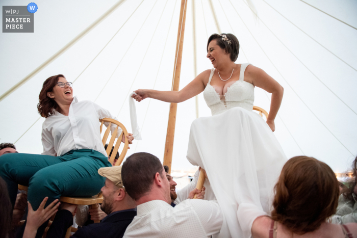 Brewster, Massachusetts winning image from the best documentary style wedding photography contest in the world of brides laughing as they are lifted up on chairs during the hora at their backyard wedding