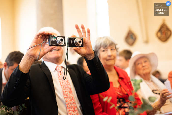 Fotografía documental de boda ganadora del concurso Isere del padre del novio sosteniendo dos cámaras tomando su foto, y una para la abuela