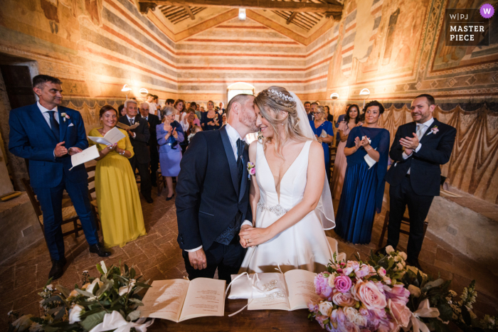 Fotografia di matrimonio documentario di livello mondiale a Siena, in Italia, presso la Chiesa di Colle Ciupi dello sposo che bacia la moglie sulla guancia durante la cerimonia