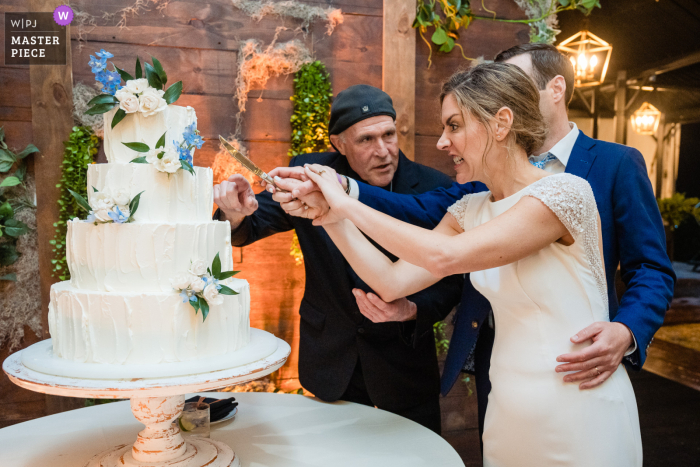 A Pennsylvania wedding photographer captured the moment when the venue coordinator guided the bride and groom in cutting their wedding cake World-class documentary wedding image with a Pennsylvania Venue coordinator showing the bride and groom how to cut the cake during a very wet day