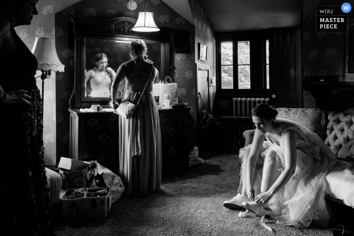 A Pennsylvania wedding photographer captured a touching moment of a mother watching her daughter change shoes and her best friend making final preparations for the introductions Philadelphia editorial style wedding photography in black and white of Mom watching on as the PA bride changes shoes for the reception and her best friend makes final preparations for the introductions