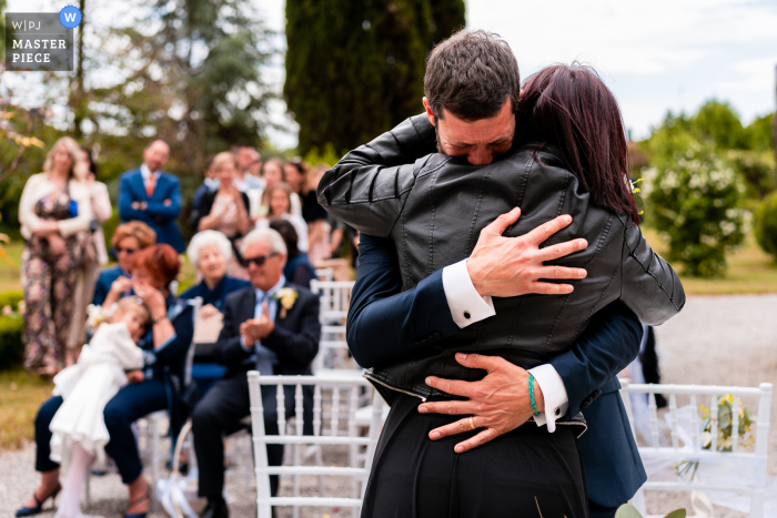 Immagine documentaristica di livello mondiale del matrimonio da Villa Iachia a Ruda, Udine, Italia che cattura un abbraccio emotivo per lo sposo fuori dalla sede