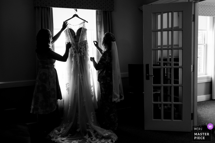 Top Maine natural wedding photography at the Southern Maine Community College created as The bride and her mom reach for her wedding gown handing in the window under outdoor lighting