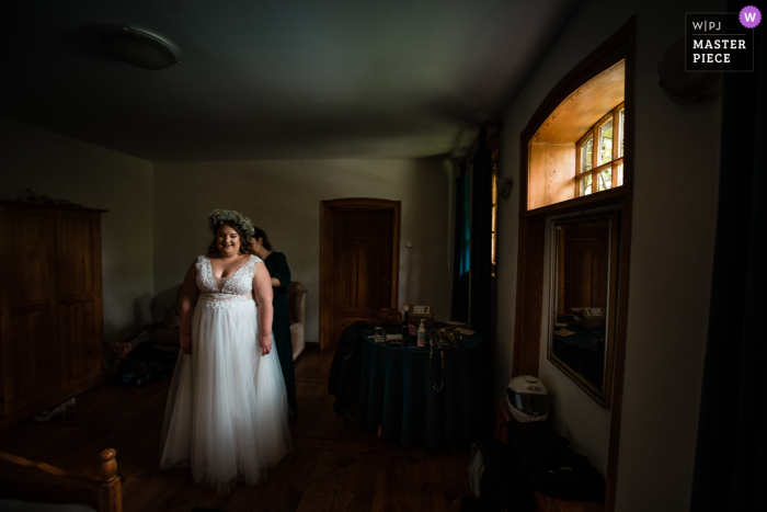 Premio a la mejor fotografía de boda estilo reportaje de Polonia en Folwark Tworzyjanki que muestra a la novia preparándose con su vestido bajo la luz de una ventana suave