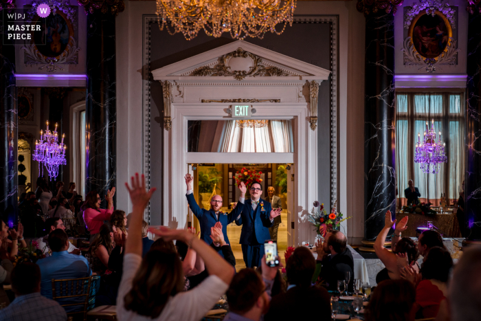 the couple enter their wedding reception as the crowd claps hands in celebration Maryland documentary wedding photography contest winning image at Hotel Monaco in Baltimore showing the couple enter their wedding reception as the crowd claps hands in celebration