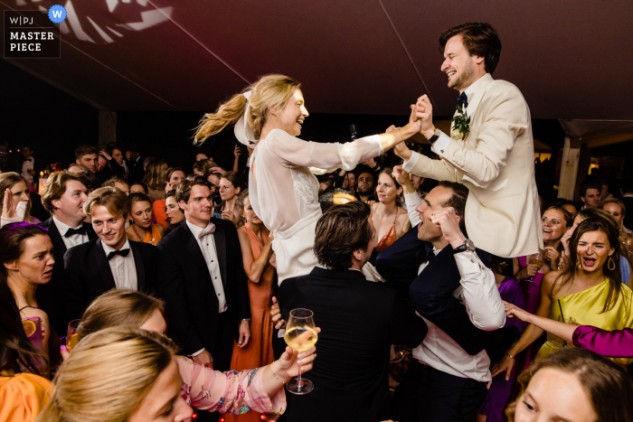 As the wedding reception gets underway at Sint Martens Laterm, the bride and groom are lifted high above the dance floor in chairs, surrounded by their guests in a joyful celebration of love