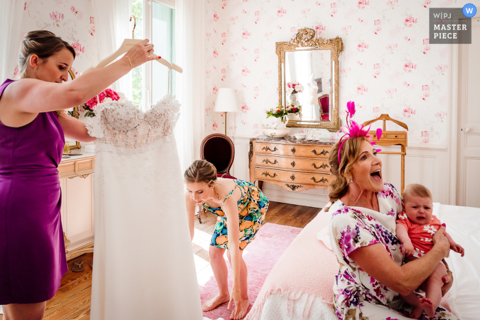 Mentre la sposa si prepara allo Chateau de Malmont in Francia, viene aiutata a stendere il vestito appeso mentre una donna siede sul letto con in braccio un bambino, catturato in un bellissimo momento dal fotografo