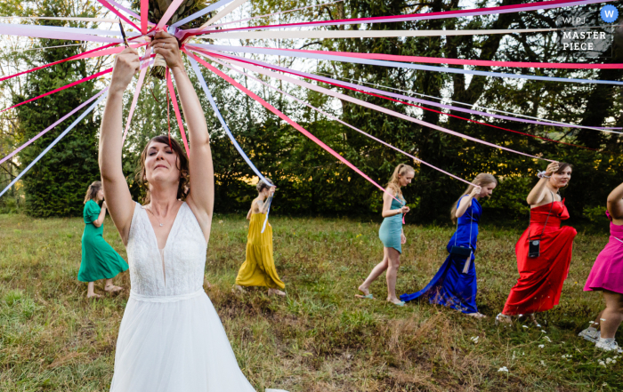 A playful wedding game on a grass lawn outdoors is captured in a photograph, featuring the bride and her bouquet's ribbons In a playful wedding game at Moulin de la Fleuristerie in Haute-Marne, the bride and bouquet's ribbons are captured in a photograph