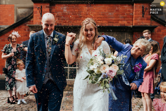 Catturata da un fotografo del Derbyshire, questa immagine di reportage di matrimonio mostra la nonna della sposa che riempie con cura il suo vestito di coriandoli mentre la coppia esce dalla Pumping House nel Derbyshire