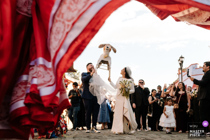 La novia y el novio capturados en una foto de boda llena de acción en la ciudad de Oaxaca, México, con una calenda de bodas brillante y un perro maché de papel blanco encima