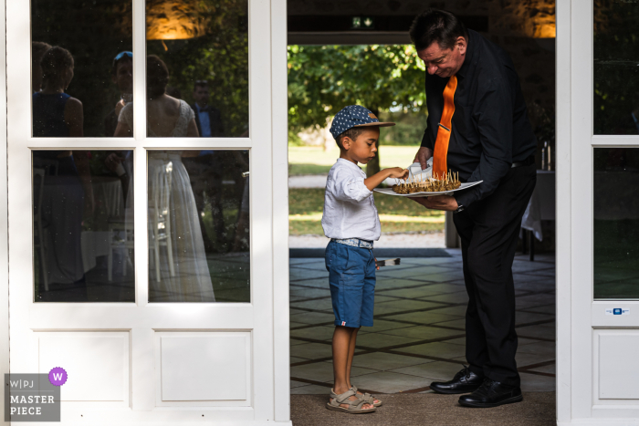 De weerspiegeling van de bruid in het vensterglas van Fleury les Aubrais, Frankrijk, werd door de fotograaf vastgelegd terwijl een jong kind hors d'oeuvres kreeg op de bruiloft