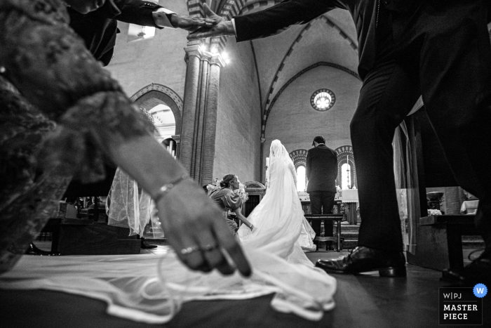 Esta fotografía de boda en blanco y negro captura el momento en que las manos ayudan a arreglar el vestido de la novia durante la ceremonia en la iglesia. Fotografía de boda de Ancona de manos ayudando a la novia a arreglar su vestido durante la ceremonia de la iglesia