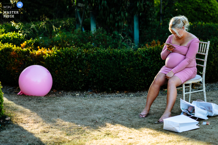 At the outdoor reception venue, a radiant pregnant wedding guest was wearing a beautiful pink dress that complemented the pink hopper toy that was next to her At the West Midlands Reception Venue, a pregnant wedding guest is wearing pink matching the pink space hopper next to her