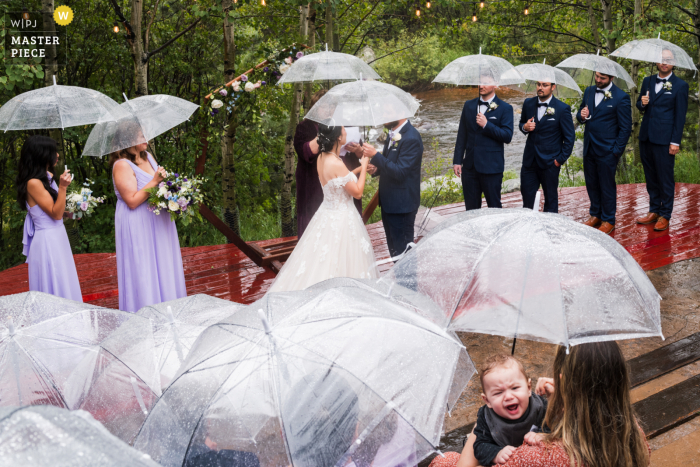 Un couple échange ses vœux au Wild Basin Lodge à Allenspark, Colorado, alors qu'une averse de pluie ajoute à l'atmosphère dramatique tandis qu'un bébé en arrière-plan ne semble pas amusé par la météo.