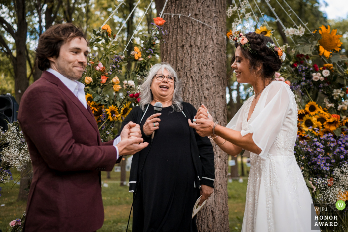 A documentary wedding photographer captured this award-winning image at a home in Lodi, Wisconsin. The bride and groom play rock, paper, scissors to decide who goes first in saying their vows, surrounded by beautiful trees.