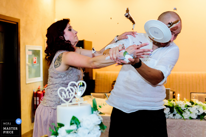 Godmother's Mischievous Cake Smash Surprises Godfather at Traditional Bulgarian Wedding in Etropole Surrounded by Bulgarian traditions, the godmother playfully surprises the godfather with a messy cake smash, adding a touch of fun to the traditional wedding in Etropole.