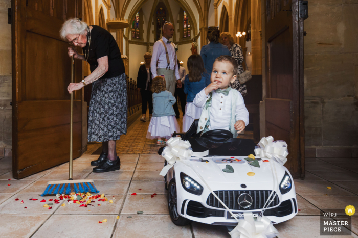 Amidst a flurry of confetti and with the help of the church caretaker, a young boy in his wedding attire takes a ride in a miniature car near the church steps.