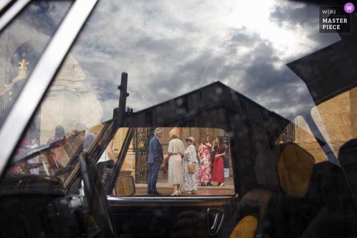 Cathedral d'Auch Frames Lively Guest Scene Through Car Window The Cathedral d'Auch provides a stunning backdrop as the car window frames a lively scene of guests mingling, their excitement palpable through the glass.
