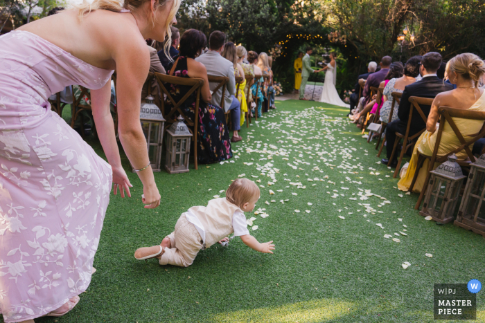 At Twin Oaks House Weddings in San Marcos, California, a curious baby makes an unexpected attempt to crawl onto the wedding aisle, bringing smiles as his mom gently reaches out to catch him.
